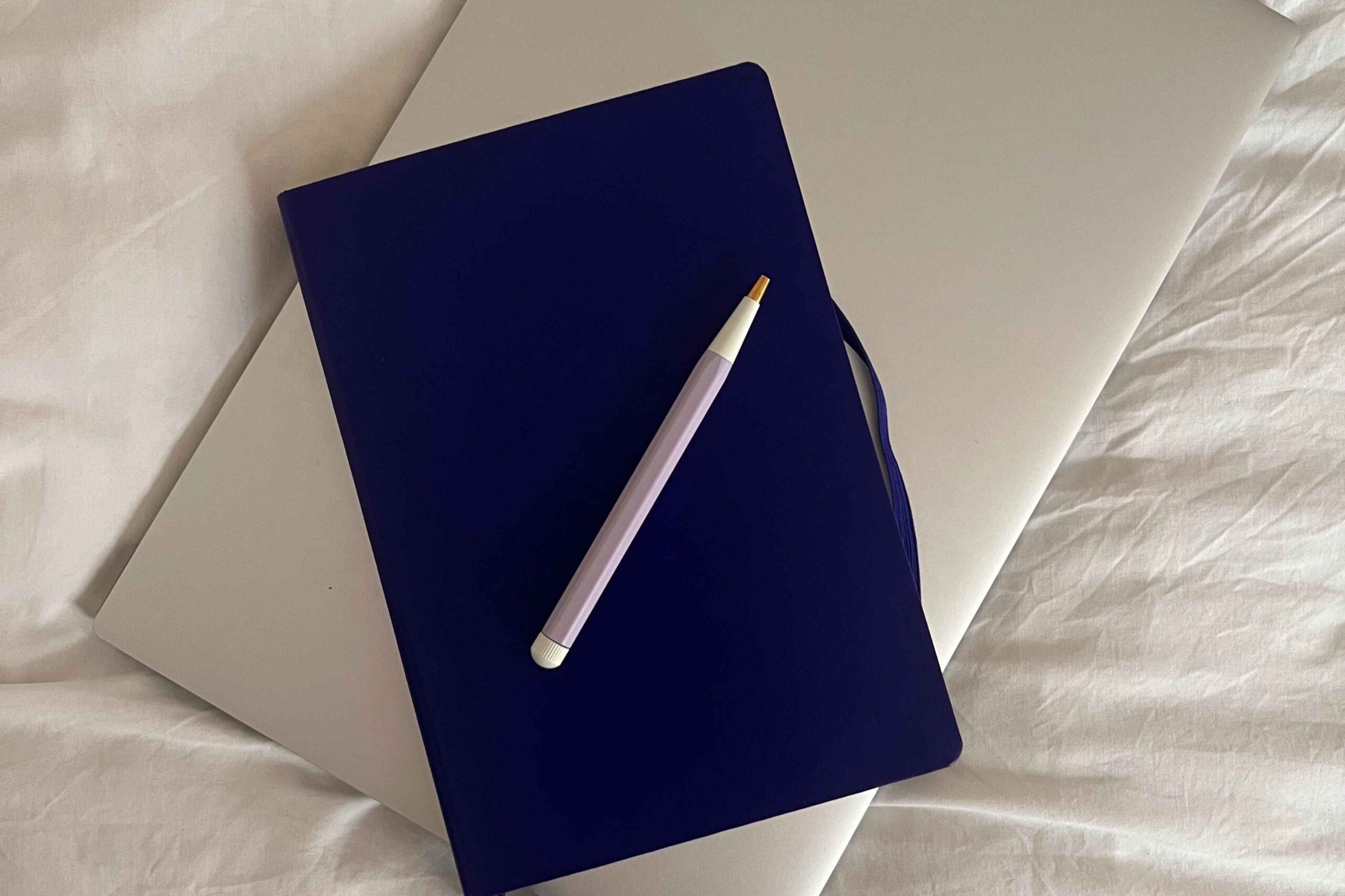 blue notebook, pen, and closed laptop stacked on top of bedsheets